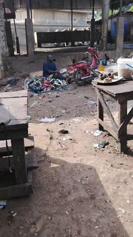The video shows an outdoor scene, likely a market or a processing area, in Awka Etiti, Nigeria. A man is sitting on the ground, dressed in a blue shirt, next to a pile of textiles. A dog is sleeping near him. In the background, there's a red moped parked. To the right, a simple wooden table holds a plate of fried fish or meat and a white bucket. Another wooden table with a blue tarp on it is visible in the foreground.

The ground is unpaved and littered with various debris and plastic waste. Chickens can be seen roaming around. In the background, under a large open-sided shelter with a corrugated roof, there are significant piles of what appear to be dried fish or similar processed goods. Other basic structures, including concrete buildings and blue barrels, are also present. The scene is brightly lit by sunlight, casting shadows and indicating it is daytime. The overall impression is that of a bustling, working environment. No visible text is present.