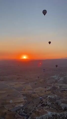 The video captures a sunrise over a valley dotted with numerous hot air balloons. The sun, a bright orange orb, is positioned low on the horizon, casting a warm glow across the sky and landscape. The sky transitions from a deep orange near the sun to a lighter shade of orange and then to a pale blue at the top of the frame. The landscape below appears rugged and somewhat arid, with valleys and hills. Several hot air balloons, in various colors and sizes, are visible at different altitudes. Some are close to the ground, while others are high in the sky. The balloons are ascending or floating, contributing to a serene and picturesque scene.