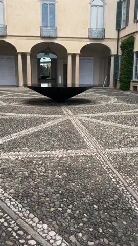 This media file captures an outdoor scene in a courtyard. The setting appears to be a historic building with arched walkways and ornate windows. The courtyard itself is paved with cobblestones arranged in a circular, radial pattern, with lighter-colored stones forming geometric lines.

Dominating the center of the courtyard is a large, flat, black, disc-like object, possibly a sculpture or a large table. It is positioned at an angle, creating a sense of depth and drawing the viewer's eye. In the background, visible through the arches, is a glimpse of what appears to be another part of the building or garden. On the walls of the courtyard, there are large posters or exhibition panels. One is partially visible on the left and another on the right, both featuring text. The text on the right-hand panel clearly reads "Josef Albers / meditations."

The time of day appears to be daylight, with soft lighting suggesting it might be overcast or late afternoon. The weather seems calm, with no visible wind or rain. The overall atmosphere is one of quiet contemplation and artistic appreciation. The scene evokes a sense of timelessness and sophisticated design, blending architectural heritage with modern art. No people are visible in the frame, emphasizing the focus on the space and the art installation. The location cues, such as the architecture and the Italian text on the exhibition panels, strongly suggest Varese, Italy, as the setting.