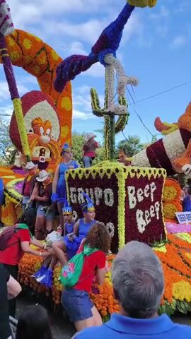 A video displays a vibrant flower parade float depicting a desert scene with cartoon characters. The float is adorned with colorful flowers, creating a detailed landscape of cacti, mountains, and a large character resembling Bugs Bunny. Several individuals, some dressed in costumes, are positioned on the float. A person in a red shirt and green scarf is visible in the foreground, interacting with someone on the float. The float also features a large sign with the word "GRUPO" and the phrase "Beep Beep" made out of flowers. The sky is partly cloudy, suggesting an outdoor event, likely a parade.