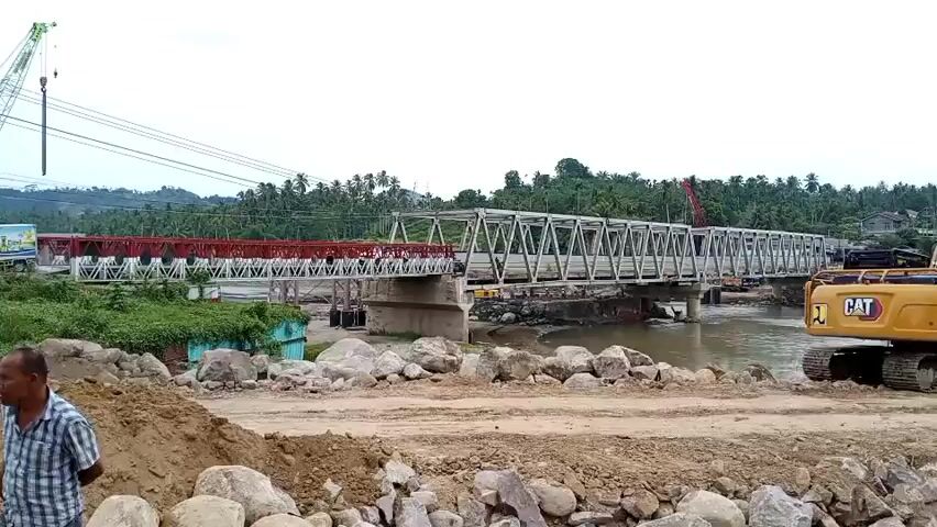 A bridge construction site is shown. The bridge appears to be a steel truss bridge, with one section painted red and white, and the other section unpainted metal. The bridge spans a river. On the right side of the frame, a yellow excavator is parked. In the foreground, piles of dirt and rocks are visible, suggesting ongoing construction work. A person in a striped shirt is partially visible on the left side of the frame. In the background, lush green trees and hills are visible under an overcast sky. A crane is also visible on the left side of the frame.