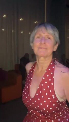 A woman with short, gray hair and tattoos on her arms is dancing and smiling. She is wearing a red halter top with white polka dots and denim shorts. The background is dark and appears to be a room with couches and curtains. The woman waves at the camera at the end of the clip.