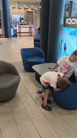 Two young children are interacting in a communal seating area with blue upholstered chairs and a small table. The child on the left, wearing a white t-shirt with a graphic print and multi-colored shorts, is kneeling and reaching into the blue armchair occupied by the other child. The child on the right, wearing a white t-shirt with a graphic print and dark pants, is leaning forward within the armchair, facing away from the camera and towards the other child. The setting appears to be an indoor public space, possibly a mall, with a display for the brand IQOS visible in the background, featuring product imagery and text. Other individuals and seating are present in the background, suggesting a commercial or waiting area.