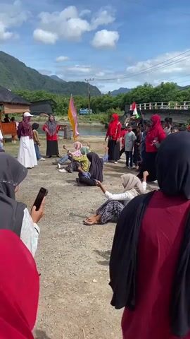 A group of people are gathered outdoors in a dirt area near a body of water. Several individuals, predominantly women and girls wearing hijabs, are participating in a game. Two participants are seated on the ground, appearing to be in a tug-of-war style competition with a rope. Others are watching, some holding phones. In the background, there are more onlookers, a bridge, mountains, and trees under a cloudy sky. Flags are visible, including one with red, white, and green colors.