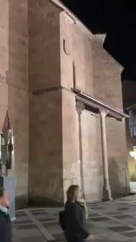 The video presents a night view of a metal sculpture situated outside a stone building.

Two women walk along the sidewalk in the foreground. A man follows behind them. They are walking away from the camera.

The sculpture, appearing to be made of polished metal, is mounted on a stone plinth. It is positioned near the corner of the building. The building is constructed from large, rectangular stone blocks, with some architectural details such as a small crest or emblem on the wall.

The lighting is dim, casting shadows from the sculpture onto the stone wall behind it. The scene suggests a quiet, urban setting at night. The focus is predominantly on the sculpture and the architectural backdrop.