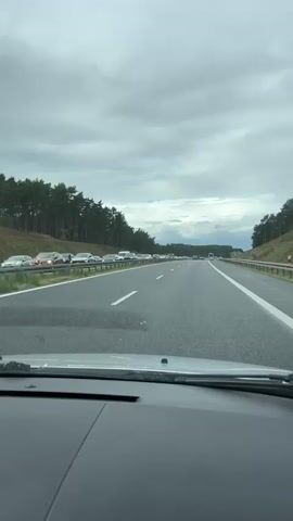 The video shows a first-person view from a car driving on a highway. The car is in the right lane of a multi-lane highway. The highway is surrounded by trees on both sides, with a grassy embankment on the right. The sky is overcast with clouds. There is traffic on the highway, with several cars visible in both lanes ahead, forming a traffic jam. The video appears to be shot during the daytime.