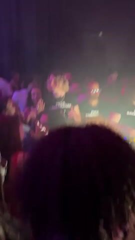 The video shows a nightclub scene. Several people are dancing and interacting with each other in a dimly lit room with pink and white lighting. In the foreground, a DJ booth features three DJs wearing matching black t-shirts. The t-shirts have white lettering. DJ equipment is visible on the booth, and some individuals are operating the equipment. Behind the DJs, a crowd of people is visible, with some dancing and others raising their hands in the air. Some faces are laughing and smiling. A neon sign in the shape of lips is visible above the crowd.