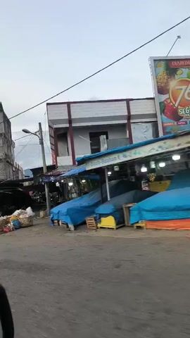 The video shows a street scene, likely in a market area, with buildings and stalls on either side. In the foreground, a car's side mirror is visible, indicating the video is shot from inside a vehicle.

The left side of the street features a multi-story building with a grey, industrial facade, and further down, several market stalls are set up with tarps and goods displayed. Some of these stalls appear to be selling produce.

The right side of the street has a more modern-looking building with a red facade and signage that reads "ROBINSON". Adjacent to this building are smaller shops and stalls covered with tarps, one of which is a vibrant blue.

The road surface is asphalt. The sky is overcast, suggesting it might be early morning or late afternoon. There are power lines visible across the sky. The overall impression is of a bustling commercial area, though the early hour or the weather might mean it's not at its peak activity.