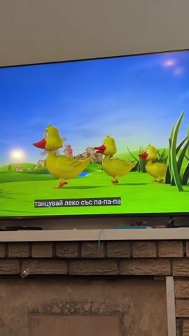 A television screen displays a cartoon video of three yellow ducklings walking across a green field. A windmill and a small village are visible in the background. The subtitles are in Bulgarian. The TV is mounted above a brick fireplace mantel. The video transitions to a close-up of one of the ducklings in a body of water, with the windmill and village in the background.