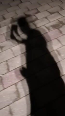 This media file is a short video clip featuring the shadow of a person walking on a wooden surface. The shadow is cast by a light source positioned above and behind the subject, creating a dark silhouette against the lighter planks. The person appears to be holding something in their right hand, possibly a phone or a small object, as their arm is raised towards their head. The movement is fluid, suggesting a casual walk. The wooden planks are arranged parallel to each other, indicating an outdoor setting like a deck or a boardwalk. The lighting suggests it is either dusk or nighttime, with an artificial light source creating the shadow. There is no visible text within the video. The location context of Nalchik, Russia, places this scene in a specific geographical and cultural setting, though the visual elements alone do not provide definitive clues to this location. The overall mood is somewhat mysterious due to the obscured nature of the subject and the dim lighting.