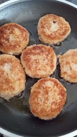 This is a close-up, top-down shot of six small, round, golden-brown patties cooking in a black frying pan with oil. The patties appear to be made of a finely ground mixture, possibly fish or chicken, and have a slightly textured surface. They are arranged in a rough circular pattern within the pan, with some slightly overlapping. The lighting is bright and appears to be coming from above, highlighting the browning of the patties and the shimmer of the oil. There is no visible action other than the cooking of the patties. The background is a dark, blurry surface that is likely the handle or edge of the frying pan.