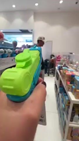 A person holds a blue and green water gun in a toy store aisle. A man in a white cap walks in the background. Shelves of toys are visible on either side of the aisle. The store is well-lit and appears clean.