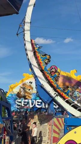 This is a video of a thrilling amusement park ride called "Seven Seas." The ride features a large pendulum-like structure that swings riders high into the air. In the foreground, people are visible standing in line or watching the ride. The ride itself is decorated with vibrant colors and imagery, including a pirate-themed design on the top. The sky is a clear blue with some scattered clouds. The ride is captured in motion, with the pendulum swinging back and forth, showing the riders experiencing the exhilaration of the ride. The video appears to be filmed during daylight hours.
