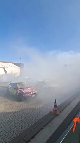 The video shows several cars performing drifts and donuts in an outdoor area. The ground is paved with cobblestones and there is a large white tent or building in the background. The sky is clear and blue. One car, which is red with various sponsorships on it, is in the foreground, creating a large cloud of smoke. There are other cars in the background also producing smoke. An orange traffic cone with white stripes is visible in the lower right corner of the frame. The scene appears to be a car show or a drifting event.