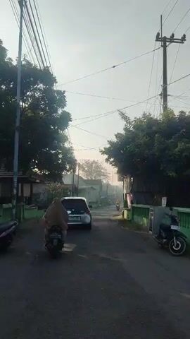 The video shows a street scene from the perspective of someone riding a motorcycle. The road is paved and narrow, with houses and trees lining both sides. Overhead, power lines crisscross the sky. In the foreground, a person in a headscarf rides a motorcycle, followed by a silver car. Further down the road, another person on a motorcycle is visible. The sun is low in the sky, casting long shadows and suggesting it is either early morning or late afternoon. The atmosphere appears calm and residential.