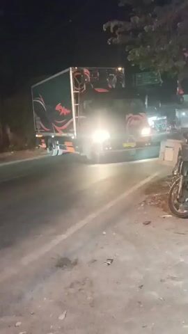 A nighttime video shows a truck with illuminated headlights driving on a road. The truck is black with orange and red graphics on the sides and the back of the cab. The graphics appear to be stylized depictions of animals, possibly dragons or birds. The truck's headlights are bright, casting a strong beam forward and illuminating the road surface, which is paved and shows some tire marks and debris. The truck's cargo box is also visible, and it appears to have some lights or equipment mounted on top. Trees and other vegetation are visible on the side of the road, as are some buildings or structures in the background. A motorcycle is parked on the right side of the road, partially in the frame. The overall scene suggests a rural or semi-urban environment at night.