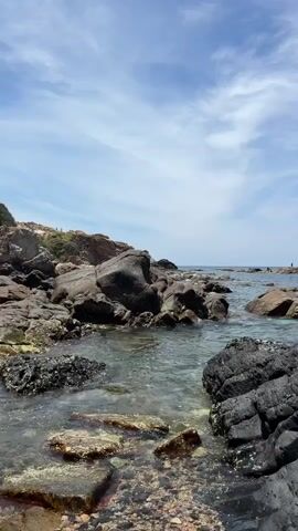 This media file depicts a rocky coastline with calm, clear water. The foreground is dominated by large, dark, textured rocks, some of which are partially submerged in the water. The water itself is shallow and clear, revealing smaller rocks and pebbles on the seabed. Beyond these rocks, the sea stretches out to the horizon, meeting a bright blue sky filled with scattered white clouds.

In the distance, several small figures can be seen standing on rocks near the water's edge, suggesting the presence of people enjoying the coastal environment. These figures are too small to discern specific details about their appearance or activities.

The scene appears to be set during the daytime, with ample sunlight illuminating the water and rocks. The sky suggests fair weather, with the clouds adding a dynamic element to the otherwise serene seascape. The presence of the sea, rocks, and the open sky points to a natural coastal location. While no specific text is visible in the media, the overall impression is one of natural beauty and tranquility.