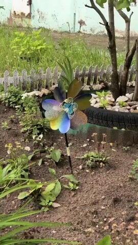 A close-up shot captures a pinwheel with colorful, petal-like blades adorned with a butterfly design in the center. The pinwheel is planted in the soil of a garden bed. The foreground is filled with rich, brown soil and various green plants, some with small white flowers. A white picket fence runs horizontally across the middle ground, separating the garden from a patch of grass and some greenery behind it. To the right, a tree trunk is visible, partially obscured by its branches and a section of a black tire, possibly used as a garden border. The background shows a portion of a building with a weathered, possibly peeling, light-colored facade. The overall scene depicts an outdoor garden setting with a decorative element.
