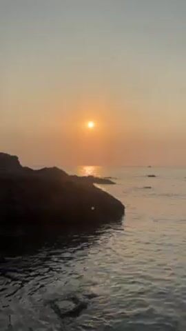 The video shows a seaside scene at sunset. A rocky promontory juts into the water in the foreground, and the sun is setting over the ocean in the background. The water is gently lapping against the rocks. It's a still shot, capturing the tranquil beauty of the sunset.