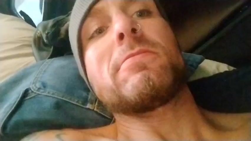 Here's a concise description of the video:

A close-up shot of a man's face. He is wearing a gray beanie and has a small ring in his nostril. He has a light beard and is lying down, appearing to be indoors. A denim jacket can be seen on or near his chest. The camera angle is looking up at his face. The lighting is a bit dark. It is a static shot with no noticeable movements or changes.