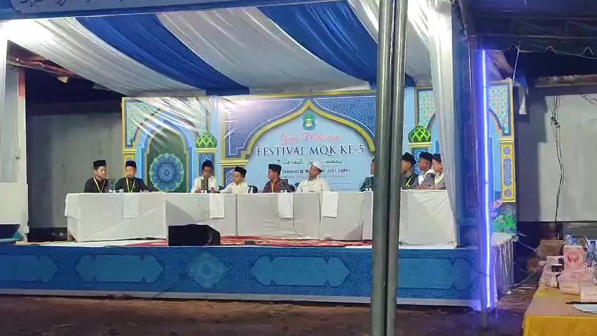 The video shows an outdoor event taking place under a tent with a blue and white striped canopy. A stage is set up with a banner in the background that reads "FESTIVAL MQK KE-5" and appears to be in Arabic script. On the stage, a group of young males, most wearing white shirts and black caps, are seated behind a long white table. They appear to be participants or judges. To the right of the stage, another table is set up with what appear to be judges or organizers, some of whom are wearing black caps. Several laptops and papers are visible on this table. The environment appears to be a community gathering or competition, possibly related to religious education given the banner's text and the attire of the individuals. There is a person in a black shirt walking away from the stage area towards the right side of the frame. The lighting is dim, suggesting it might be evening or nighttime.