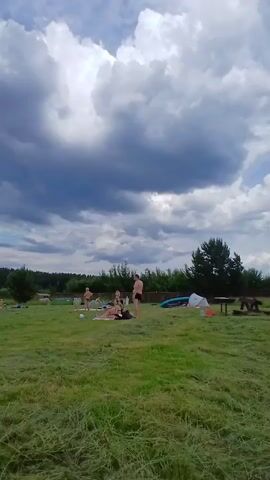 A group of people are visible on a grassy area with a lake and trees in the background. The sky is cloudy. In the foreground, on the grass, there are several objects, including what appears to be a hookah pipe and some shoes. In the middle ground, several people are scattered around. Some are sitting or lying on the grass, while others are standing. One person is walking towards the left of the frame, and another is walking towards the right. Further in the background, near the lake, more people are visible, some of whom appear to be in the water. There are also some structures along the shoreline, including what looks like a pier or dock. A large tree is prominent on the left side of the frame. The overall scene appears to be a relaxed outdoor gathering or beach area.