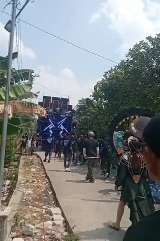 The video captures an outdoor procession or parade. In the background, a large sound system with multiple speakers and flashing lights is visible, suggesting a musical event or party. A group of people dressed in dark clothing and masks walks in a line towards the sound system. In the foreground, a person in an elaborate black costume with a large circular headpiece that resembles a mouth with teeth is walking away from the camera. Another person in a light-colored jacket is walking towards the procession, and a third person in a black t-shirt with white and blue lettering is partially visible in the lower right corner. The environment appears to be a rural or suburban street, with trees and vegetation on either side of the path. The ground is paved, and there are some debris and construction materials along the side of the path. The sky is blue and clear, indicating a daytime event.