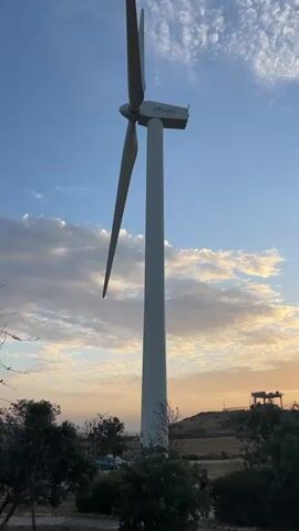 The video shows a tall white wind turbine with the name "alcon" printed on it, set against a blue sky with wispy clouds and a sunset glow. The camera pans right to show more wind turbines in the distance, partially obscured by trees and shrubs. The landscape includes a field and a structure on a hill. The sun is setting, casting a warm light on the scene.