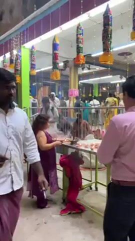 People are gathered indoors, tending to small lit oil lamps arranged on a table. Many are dressed in casual clothing; others are wearing traditional attire. Overhead hangs an array of colorful decorations. The room is well-lit with bright lights. Some smoke rises from the lamps, obscuring the view in places. A metal railing separates the people and the lamps. Some people are interacting with each other, while others focus on the lamps. The general impression is one of a religious ceremony or offering.