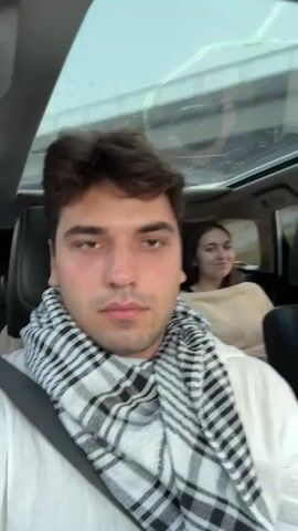 The video shows four people in a car. The person in the foreground, wearing a white shirt and a black and white checkered scarf, is looking at the camera and smiling. Behind them, a man in a light-colored t-shirt is visible, looking to his right. Further back, two women are seated. The woman on the left, wearing a light-colored top and a checkered scarf, is smiling and looking towards the camera. The woman on the right is also wearing a checkered scarf and a light-colored coat, and is looking away from the camera. The car has a sunroof through which the sky and trees are visible. The scene appears to be shot during the daytime.