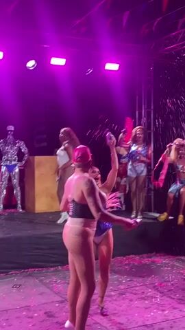 A group of people are on a stage. The stage is decorated with pink and purple lights. There are confetti and streamers on the stage. In the foreground, two people are dancing. One person is wearing a pink outfit and a pink hat. The other person is wearing a blue outfit. In the background, there are other people on the stage, some of whom are holding flags and props. The audience is not visible.