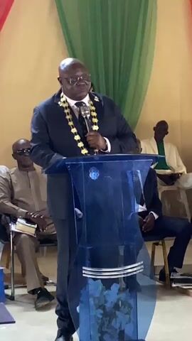 A man in a dark suit and a gold chain stands at a blue podium, speaking into a microphone. He is wearing glasses and has a bald head. In the background, several other people are seated, some of whom are wearing suits. The room is decorated with green and red fabric draped from the ceiling. The man appears to be giving a speech or presentation.