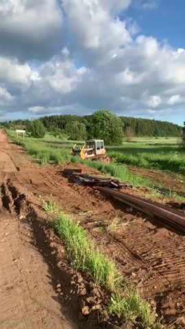 The video begins with a view of a dirt road under a cloudy sky. A yellow bulldozer is visible in the distance, situated in a grassy field. The road appears to be under construction or repair, with tire tracks and loose dirt visible. As the camera moves, it shows piles of pipes and other construction materials. The road continues through a rural landscape with trees and fields. Another yellow excavator is seen in a field on the right side of the road. The camera pans to show houses and more trees in the background. The video concludes with a shot of the road and the surrounding landscape under the same cloudy sky.