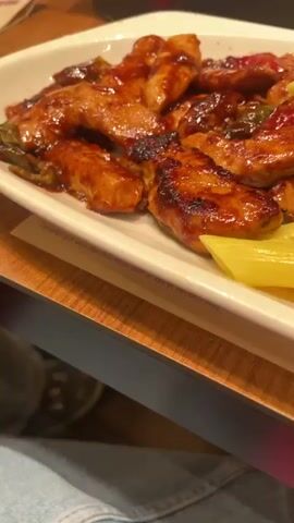 The media features a close-up, eye-level shot of a white rectangular plate filled with food. The plate is positioned on a wooden surface, likely a table. The lighting suggests an indoor setting, possibly a restaurant or home dining area, with no clear indication of the time of day or weather.

The food on the plate consists of two main components. On the left side, there are pieces of glazed chicken, appearing to be stir-fried with vegetables such as red peppers and green chilies, all coated in a dark, glossy sauce. This section of the plate is closer to the camera, with the chicken pieces prominently displayed.

To the right of the chicken is a generous portion of penne pasta. The pasta is a light yellow color and appears to be seasoned with green herbs, possibly parsley or oregano. The pasta is arranged in a pile, filling the right side of the plate.

Further to the right and towards the bottom of the plate is a small salad. The salad includes lettuce, shredded carrots, and a single cherry tomato. A hint of salad dressing can be seen on the edge of the plate.

In the background, out of focus, are elements of the setting. To the left, a portion of a person's leg, clad in blue denim jeans, is visible, indicating a person is seated at the table. Behind the plate, on the table, are indistinct objects that could be condiments, napkins, or other restaurant tableware, along with a blurred view of what looks like packaging or menu items, suggesting a dining establishment. The overall impression is of a meal being served.

There is text visible on the table's surface underneath the plate, which appears to be a placemat or menu with a QR code printed on it. Another piece of text, "BAGLI BASI," is partially visible on the placemat. The context of "Bağcılar, Turkey" indicates a likely geographical location.
