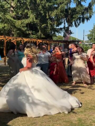 A blonde bride in a large white wedding dress with intricate lace detailing on the bodice is shown from behind, with her back to the camera. She is wearing a tiara and holding a bouquet of flowers. She is standing on a grassy lawn in what appears to be an outdoor venue, with a wooden pergola and trees in the background. She is facing a group of women and a few children, who are also on the lawn. The women are dressed in various colored dresses, and they appear to be anticipating something. One woman in an orange dress and another in a pink dress are reaching up, possibly to catch something. The overall atmosphere is celebratory and appears to be a wedding reception.
