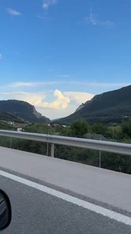 A video captures a view from a moving vehicle, looking out towards a rural landscape under a partly cloudy blue sky. In the foreground, the edge of a road with white lane markings is visible, separated from the environment by a silver metal guardrail. Beyond the guardrail, there are trees and vegetation. In the mid-ground, a valley opens up between rolling, tree-covered mountains. Some buildings, possibly houses, are scattered throughout the valley. A bright white cloud formation is prominent in the sky above the valley. A single traffic cone with red and white stripes sits on the side of the road. The camera appears to be moving from left to right, following the curve of the road.