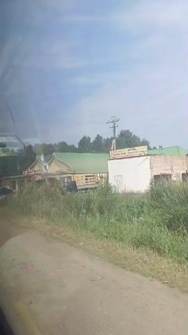 A video shows a building with a green roof and wooden exterior. The building has a sign that reads "Встреча" which translates to "Meeting" or "Encounter." There are several cars parked in front of the building, including a white sedan, a dark sedan, and a dark van with a flatbed trailer. The building appears to be a cafe or restaurant. There is also a tall utility pole with wires. The area around the building is covered in grass and trees. The sky is cloudy. The video is taken from a moving vehicle.