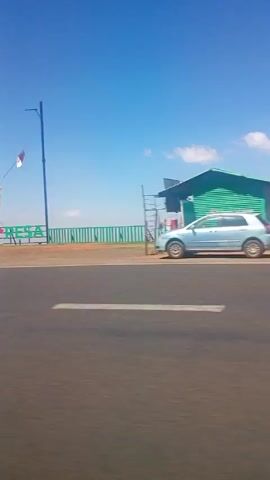 The video shows a roadside view. First, a large sign for "M-PESA" is seen, with a hand holding a cell phone. Next, the video shows some small buildings on the side of the road, possibly shops or stalls. Then a truck filled with items drives by. Finally, the video shows a landscape of red rock and vegetation alongside the highway. The sky is blue throughout.