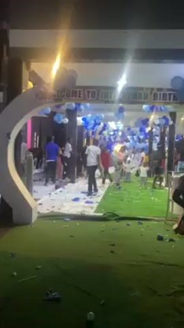 Here is a forensic, matter-of-fact description of the scene in the video:

**Setting:** The video is shot outdoors at night, with artificial lighting illuminating a party venue. The foreground is covered in green artificial turf.

**People:** There are several people visible, including children and adults. They are dressed in various casual and party attire. A young boy is standing in the foreground of the video, looking to the right.

**Objects:** A sign reads, "WELCOME TO JOE JONATHAN BIRTHDAY PARTY." The entrance is decorated with white and blue balloons and white streamers. Tables and chairs are set up throughout the area. Scattered confetti and streamers are on the ground.

**Actions:** Some people are standing around, while others are walking through the party area. There appears to be a celebration in progress, with confetti scattered.

**Interactions:** People are interacting with each other and moving around the party space. There is a sense of activity and celebration.

**Environment:** The environment appears to be a designated outdoor party area with artificial turf, decorated specifically for a birthday celebration. The overall atmosphere is festive.