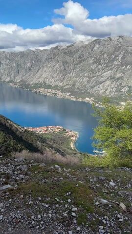 The video shows a scenic view of a bay surrounded by mountains. The water is a deep blue color and reflects the sky. A small village with red-roofed houses sits on a peninsula jutting out into the bay. The mountains are gray and rocky, with some patches of green vegetation. The sky is partly cloudy with patches of blue. The foreground shows a rocky hillside covered with moss and sparse vegetation. A green tree branch extends into the frame on the right. The camera slowly zooms in on the village.