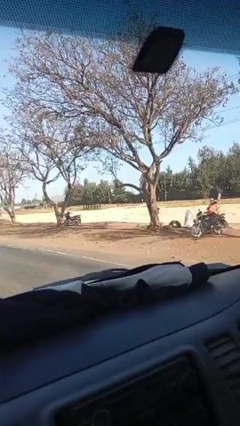 The video shows a view from inside a vehicle driving on an asphalt road on a sunny day. On the right side of the road, there is a sandy area with two bare trees casting shadows. Two motorcycles are parked in the shade of the trees. A few people are walking around in the sandy area and further in the background, there are more trees and some buildings. The foreground shows the dashboard of the vehicle.