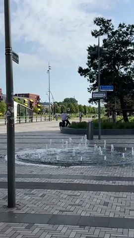 The video captures a public space in what appears to be a European city on a sunny day. In the foreground, there is a circular fountain made of concentric rings, with small jets of water spraying upwards. The ground around the fountain is paved with grey rectangular bricks. A man on a bicycle with panniers is visible in the background, stationary near some shrubbery and a trash receptacle. Beyond him is a white modern bus shelter, a street, and red brick buildings with yellow awnings. Street signs can be seen on a lamppost and a tree. The sky is blue with some clouds.