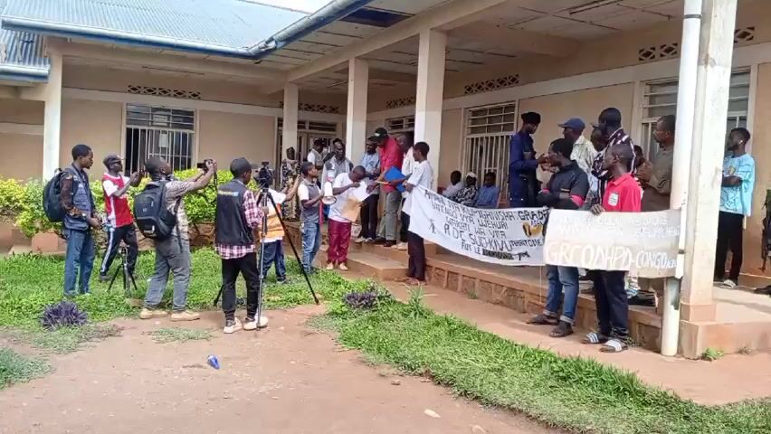 The video shows a group of people gathered in front of a building with a porch, possibly for a press conference or demonstration. Several people are holding cameras and recording the event. A man wearing a red shirt and a baseball cap is standing on the porch, holding a microphone, and reading from a document. Another person is also holding a microphone. Several people are holding a banner with text written on it. Another person is holding a sign with text. The building appears to be a one-story structure with multiple windows and white columns supporting the porch roof. The environment suggests a warm climate with green grass and vegetation visible.