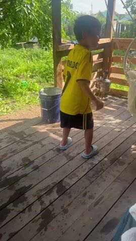 A person's foot wearing a white clog is visible in the foreground, resting on a green chair. In the background, a young child wearing a yellow t-shirt with the name "RONALDO" and the number "7" on it, and black shorts, stands on a wooden deck. The child is holding a thin rod. To the left, a blue bucket is visible. A samovar and other objects are on the deck behind the child. The deck is wet, with water stains visible. Green grass and foliage are in the background.