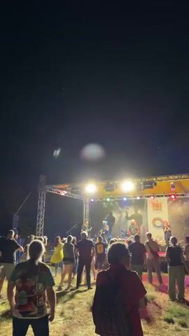 This is a vertical, eye-level, outdoor video capturing a nighttime outdoor concert. A band is performing on a stage, which is illuminated by bright stage lights and a backdrop banner. The stage is constructed with metal trusses. The crowd is gathered in front of the stage, with many people visible from the back, looking towards the performers.

The band consists of at least four members: a guitarist on the left, a vocalist/musician in the center wearing sunglasses and playing a guitar, a drummer on the far right, and another musician to the right of the central figure. The mood appears to be energetic and engaged, with audience members seemingly enjoying the music.

The background is dark, indicating it's nighttime. The ground where the audience stands is covered in dry grass or straw. The setting is likely a public space or festival grounds. The weather appears to be clear, as there are no signs of rain or wind.

One visible logo on the stage backdrop is circular and features stylized imagery, possibly representing a local event or festival. Another graphic on the banner includes text that is difficult to decipher clearly due to the lighting and resolution. However, one distinct text element visible on the banner is a colorful graphic with a guitar and other musical symbols, alongside a circular emblem.

The time of day is clearly night, indicated by the dark sky and artificial lighting of the stage. The location is identified as Tutrakan, Bulgaria. The overall scene is one of a lively outdoor music performance at night.