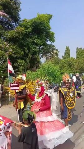 A parade is underway with participants dressed in elaborate costumes. In the foreground, a young person in a black shirt holds a long stick with a colorful lantern attached. To their right, a person in a large, ornate red and white costume with feathery white wings stands prominently. Behind them, several individuals wear distinct, theatrical costumes. One person is adorned in a black and gold outfit with a large mask featuring horns and elaborate headwear. Another person sports a black garment with colorful, sequined accents and bright orange footwear. Further down the street, more participants in similar dark, decorated attire are visible, forming a procession. The parade is taking place outdoors on a paved road, bordered by lush green trees and foliage. A few motorcycles are parked on the side of the road. The sky is clear and blue, suggesting a sunny day. A small Indonesian flag is visible on a pole held by one of the parade participants. The overall atmosphere appears festive.