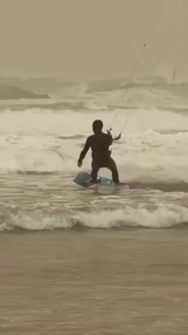 A lone kitesurfer navigates turbulent waves, visible from the back, clad in a dark suit, holding onto the kite's control bar. The person maneuvers through choppy water, where large waves break in the background. The sky is overcast, creating a monochromatic atmosphere. A transmission tower is visible in the distance along the horizon.