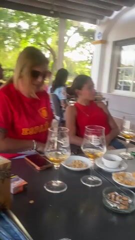 The video shows a group of people sitting at an outdoor cafe table. A woman in a red shirt with the Spanish flag on it is on the left, smiling and clapping. A woman in a red top is in the center, looking at the camera and vaping. Two glasses of beer and bowls of nuts are on the table, along with an ashtray full of cigarette butts and a can of Coca-Cola. A television screen mounted overhead displays a soccer match. In the background, other patrons are visible, and trees and buildings can be seen.