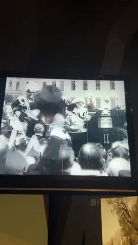 This is a black and white video clip playing on a screen, possibly in a museum or exhibition. The video appears to be historical footage. The first few seconds show a blurred, chaotic scene with many people in hats, suggesting a crowd or gathering. Then, the scene shifts to a more focused view of a man on horseback, dressed in elaborate military-style attire and a tall hat. He is surrounded by several other individuals, also in uniforms and hats, who appear to be attending to him and the horse. A carriage or cart is partially visible behind the horse. The background suggests an outdoor setting, possibly a park or field, with trees and some buildings in the distance. The overall impression is of a formal event or procession from a bygone era.