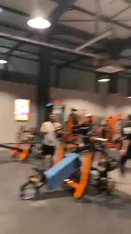 A static, medium shot captures the interior of a busy gym. Numerous individuals are spread out across the large space, engaging with various weightlifting machines, identifiable by their bright orange color. The gym is well-lit with multiple hanging light fixtures, and the ceiling has an industrial, truss-like structure. In the foreground, a barbell rests on a rack. The floor is a neutral gray, and the walls are a muted gray color. The gym appears well-equipped and is actively being used by several people.